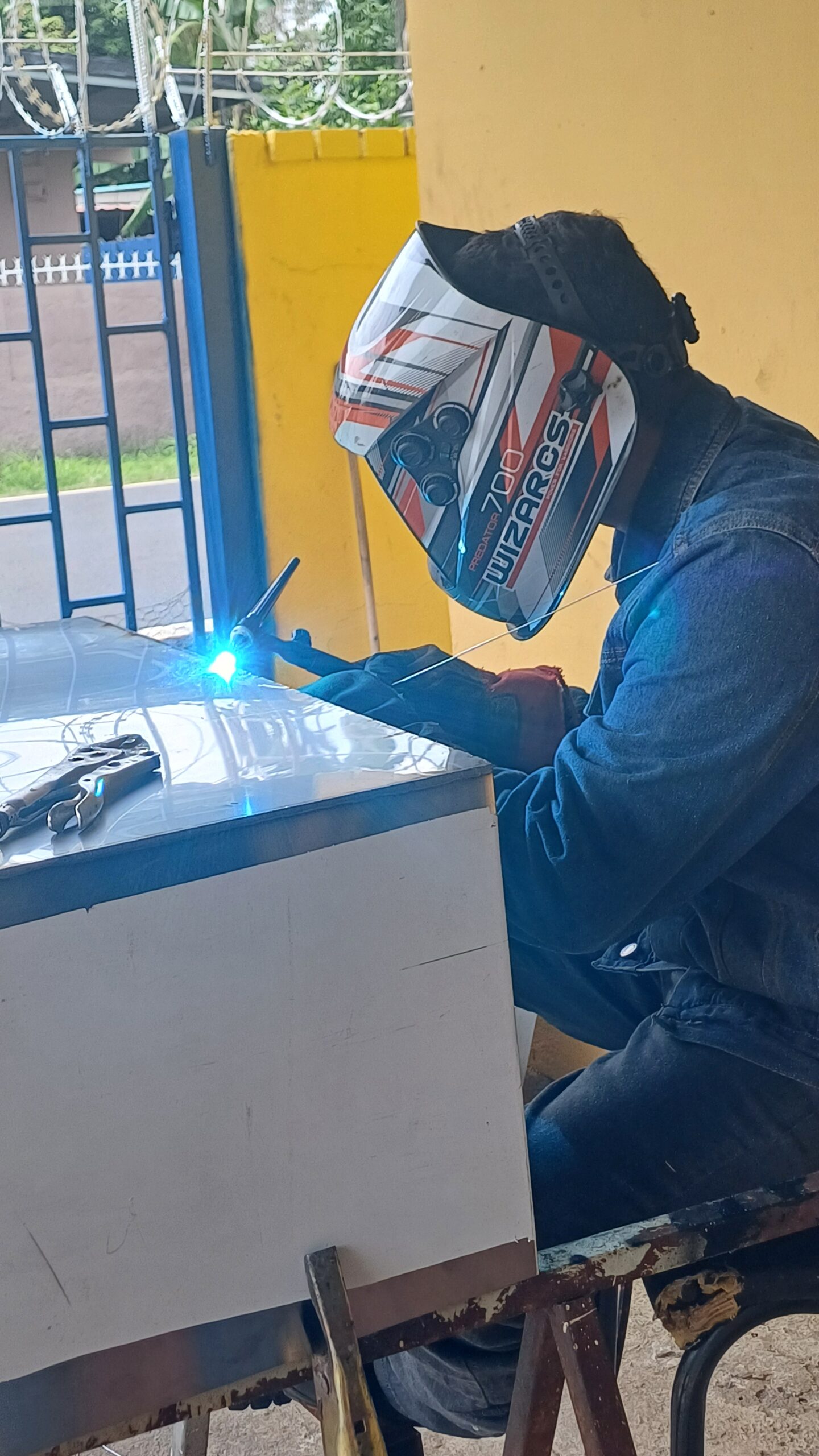 Un operario con careta de soldar blanca y naranja modelo "Predator 700 Wizarcs", camisa de mezclilla y guantes de protección realiza una soldadura por arco eléctrico sobre una mesa metálica, generando un destello de luz brillante.