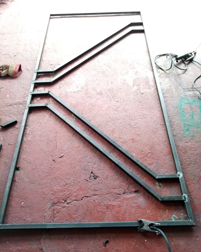 Un marco rectangular de metal negro con un diseño interior geométrico y asimétrico de barras diagonales soldadas yace en el suelo de concreto de color terracota. Cerca hay guantes de trabajo y herramientas.
