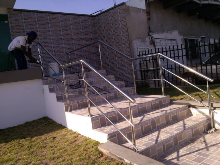 Escalinata exterior revestida con baldosas cerámicas texturizadas, equipada con barandas dobles de acero inoxidable a ambos lados, mientras un trabajador ajusta detalles en la parte superior.
