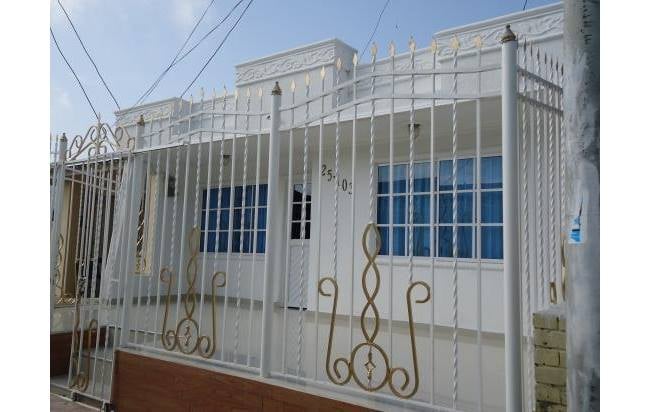 La fachada de una casa de una planta pintada de blanco está protegida por una reja de seguridad metálica blanca que cubre toda la parte frontal. La reja tiene barrotes verticales y paneles decorativos con volutas de hierro forjado pintadas de color dorado. En la pared de la casa hay un letrero con el número "25-01". Las ventanas tienen persianas azules. La base de la reja está rematada con un zócalo de imitación madera.