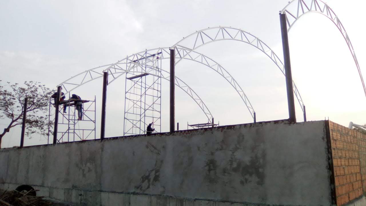 Vista exterior de una obra donde se levanta una estructura de arcos metálicos sobre muros de concreto y ladrillo. Dos operarios trabajan en un andamio en el lateral izquierdo.
