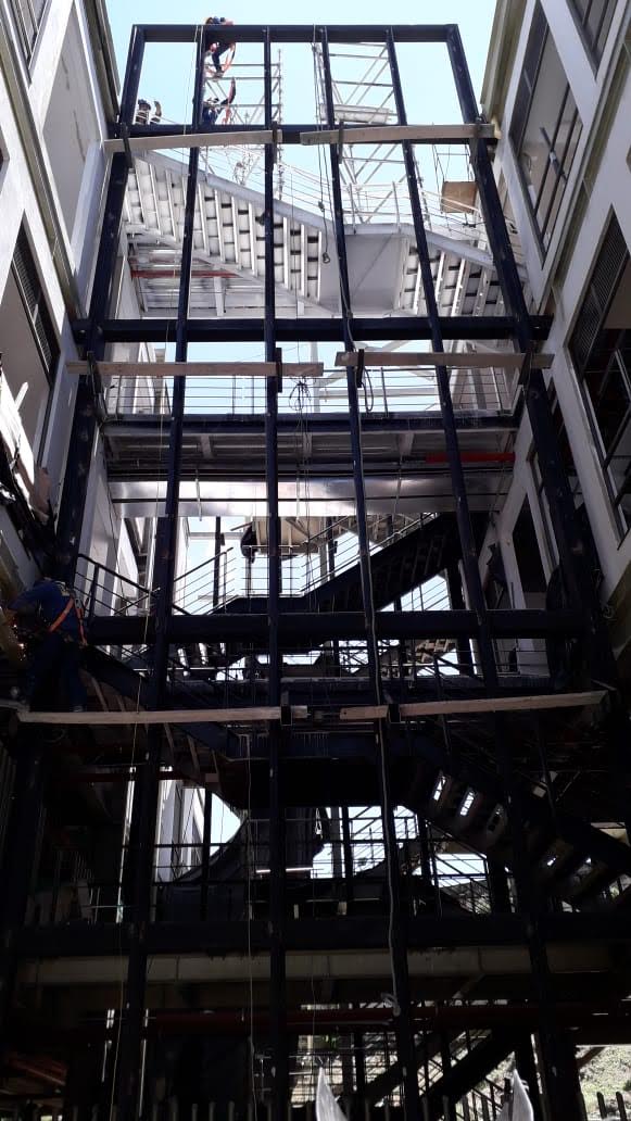 Vista en contrapicado de una compleja estructura de acero oscura en el interior de un edificio, con múltiples niveles, escaleras metálicas y andamios donde trabajan operarios, bajo una cubierta superior abierta.