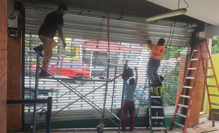 Tres operarios en un garaje trabajando en la instalación y ajuste de un portón enrollable de láminas metálicas. Utilizan un andamio móvil y una escalera de tijera, con herramientas y correas en el suelo.