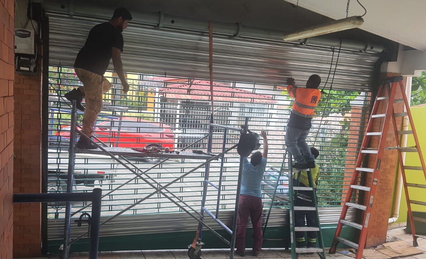 Tres operarios en un garaje trabajando en la instalación y ajuste de un portón enrollable de láminas metálicas. Utilizan un andamio móvil y una escalera de tijera, con herramientas y correas en el suelo.