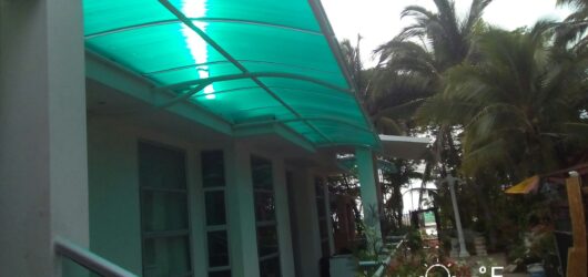 Vista exterior de una estructura de cubierta con techo arqueado de color verde translúcido, sostenida por columnas blancas. Está adosada a una casa moderna con grandes ventanales y se ubica en un jardín con palmeras y vegetación. Incluye una marca de agua de fecha y temperatura.
