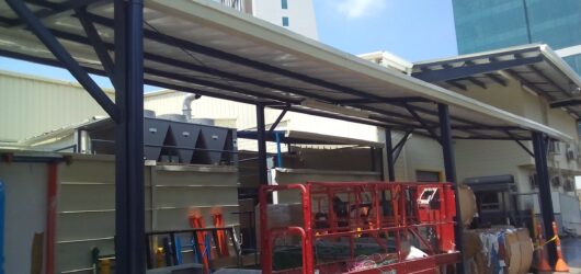 Vista exterior de una cubierta metálica de color oscuro con techo traslúcido, sostenida por columnas robustas. Protege una zona de carga y servicio donde se observa una plataforma elevadora de tijera de color rojo brillante, equipos industriales y material de embalaje en el suelo. Al fondo se aprecian edificios altos modernos bajo un cielo azul.