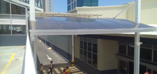 Vista desde una rampa de acceso elevada que mira hacia abajo a una rampa peatonal exterior de concreto. La vía está protegida por una cubierta metálica con techo de policarbonato curvo oscuro, sostenido por columnas blancas. Tres personas, incluyendo un empujador de carrito de compras, caminan por la rampa. Al fondo se ven edificios modernos de gran altura y un cielo azul claro.