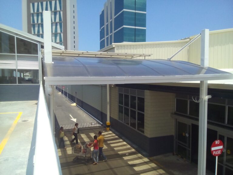 Vista desde una rampa de acceso elevada que mira hacia abajo a una rampa peatonal exterior de concreto. La vía está protegida por una cubierta metálica con techo de policarbonato curvo oscuro, sostenido por columnas blancas. Tres personas, incluyendo un empujador de carrito de compras, caminan por la rampa. Al fondo se ven edificios modernos de gran altura y un cielo azul claro.