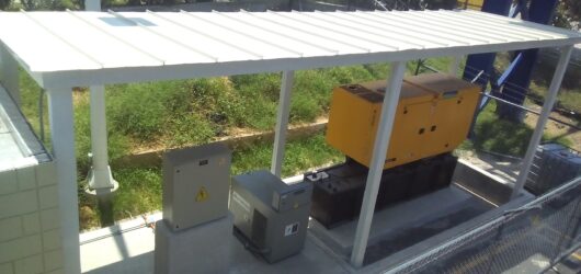 Vista superior de una cubierta metálica plana de color blanco, sostenida por delgadas columnas que protegen equipos eléctricos y un generador amarillo al aire libre. La estructura se ubica en un área cercada con malla ciclónica, vegetación alta y edificios de fondo.