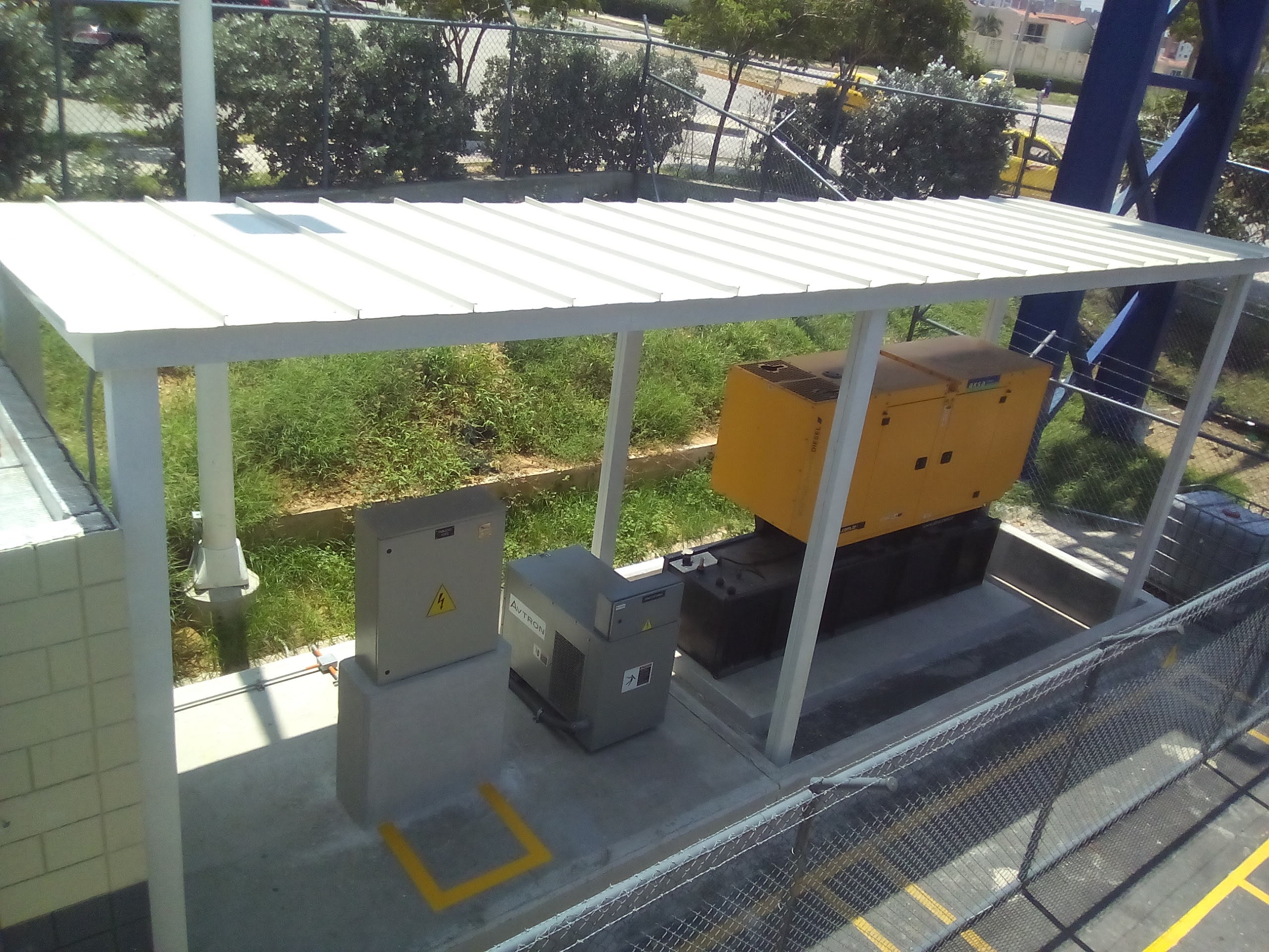 Vista superior de una cubierta metálica de color blanco con diseño plano e industrial, sostenida por delgadas columnas que protegen equipos eléctricos y un generador amarillo al aire libre.