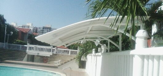 Vista exterior de una estructura metálica con techo arqueado de color blanco situada al lado de una piscina, rodeada por muros blancos y palmeras bajo un cielo despejado.