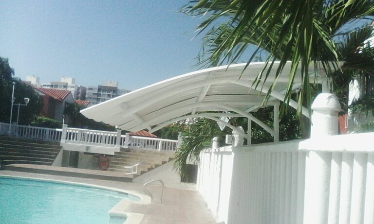 Vista exterior de una estructura metálica con techo arqueado de color blanco situada al lado de una piscina, rodeada por muros blancos y palmeras bajo un cielo despejado.