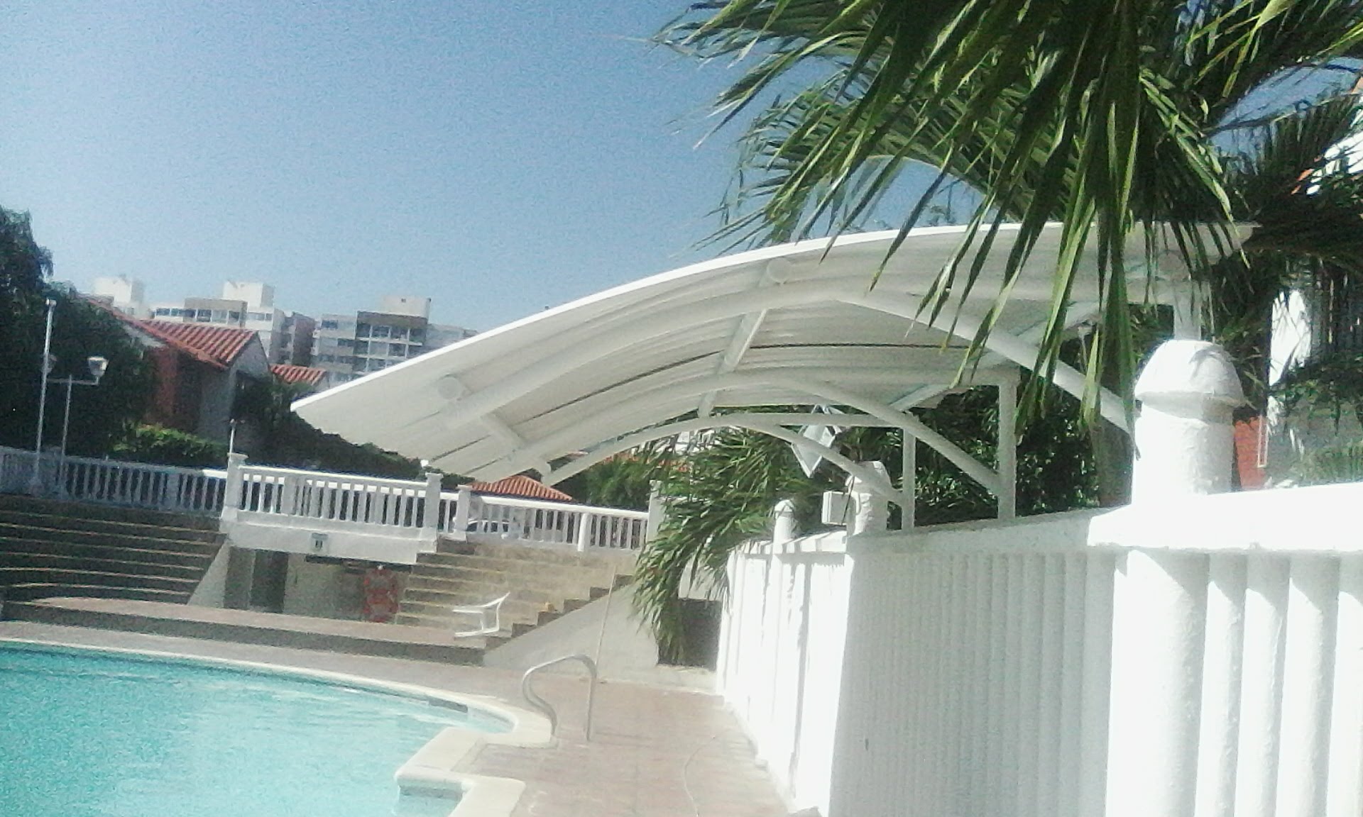Vista exterior de una estructura metálica con techo arqueado de color blanco situada al lado de una piscina, rodeada por muros blancos y palmeras bajo un cielo despejado.
