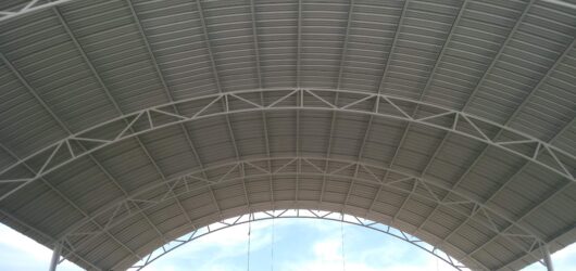 Vista inferior de una gran cubierta metálica con forma de arco, mostrando las láminas de zinc corrugado y el entramado de vigas y cerchas de acero blanco que la sostienen, con un cielo parcial de fondo.