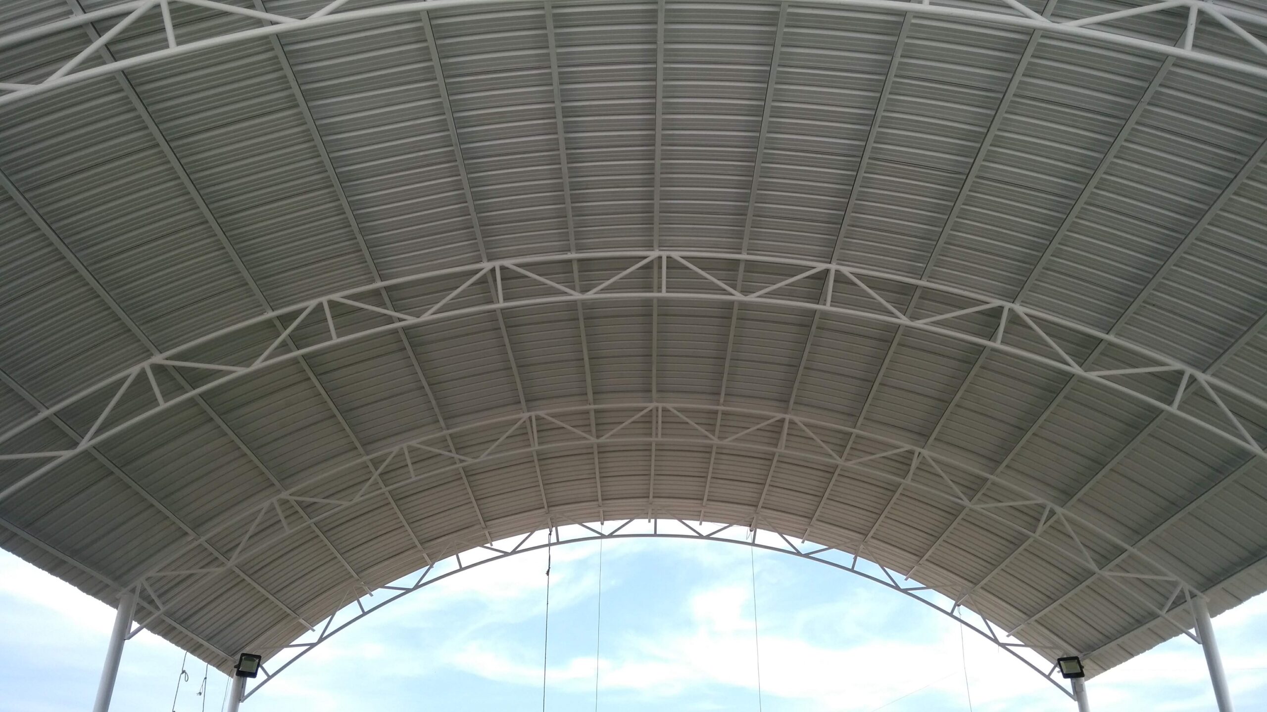 Vista inferior de una gran cubierta metálica con forma de arco, mostrando las láminas de zinc corrugado y el entramado de vigas y cerchas de acero blanco que la sostienen, con un cielo parcial de fondo.