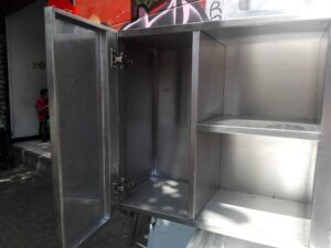 Primer plano de un gabinete colgante de acero inoxidable abierto. La puerta izquierda está abierta hacia afuera mostrando bisagras metálicas ajustables. El interior cuenta con una división central y un estante horizontal. Se observa una persona sentada al fondo en un local comercial.