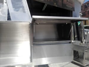Un módulo de mueble de acero inoxidable con una puerta superior horizontal abierta mediante pistones hidráulicos metálicos. Abajo se aprecia un panel frontal liso y, a la derecha, una pequeña estructura auxiliar de acero. Al fondo, la fachada del taller con un letrero comercial.