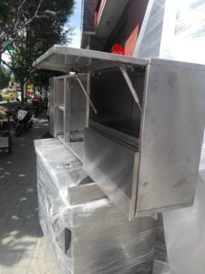 Un conjunto de gabinetes y muebles de acero inoxidable apilados en la acera frente a un taller. Un compartimento superior permanece abierto con pistones hidráulicos, mostrando su espacio interno. En la calle transitan vehículos y se aprecian árboles y edificaciones al fondo.