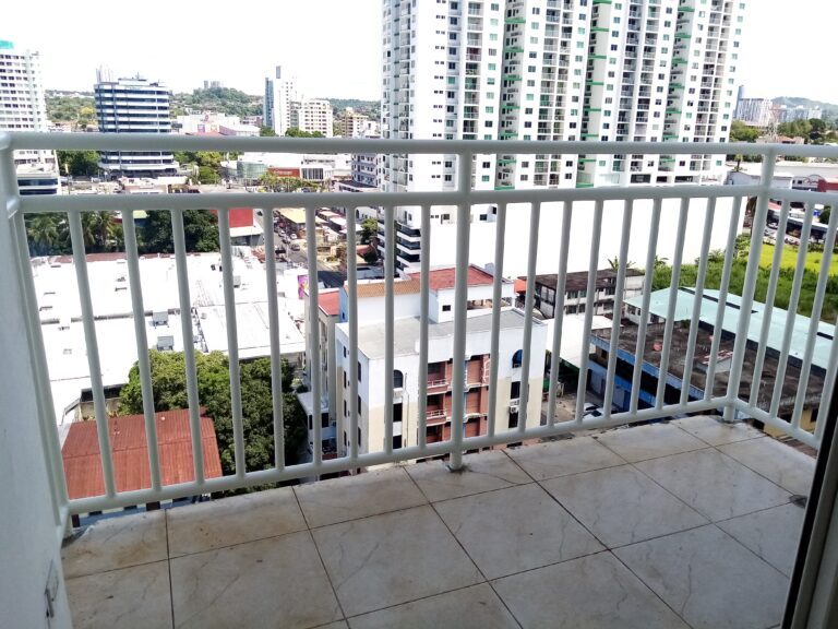 Perspectiva amplia desde un balcón con suelo de baldosas y baranda blanca, mostrando torres de apartamentos y construcciones comerciales en el horizonte.