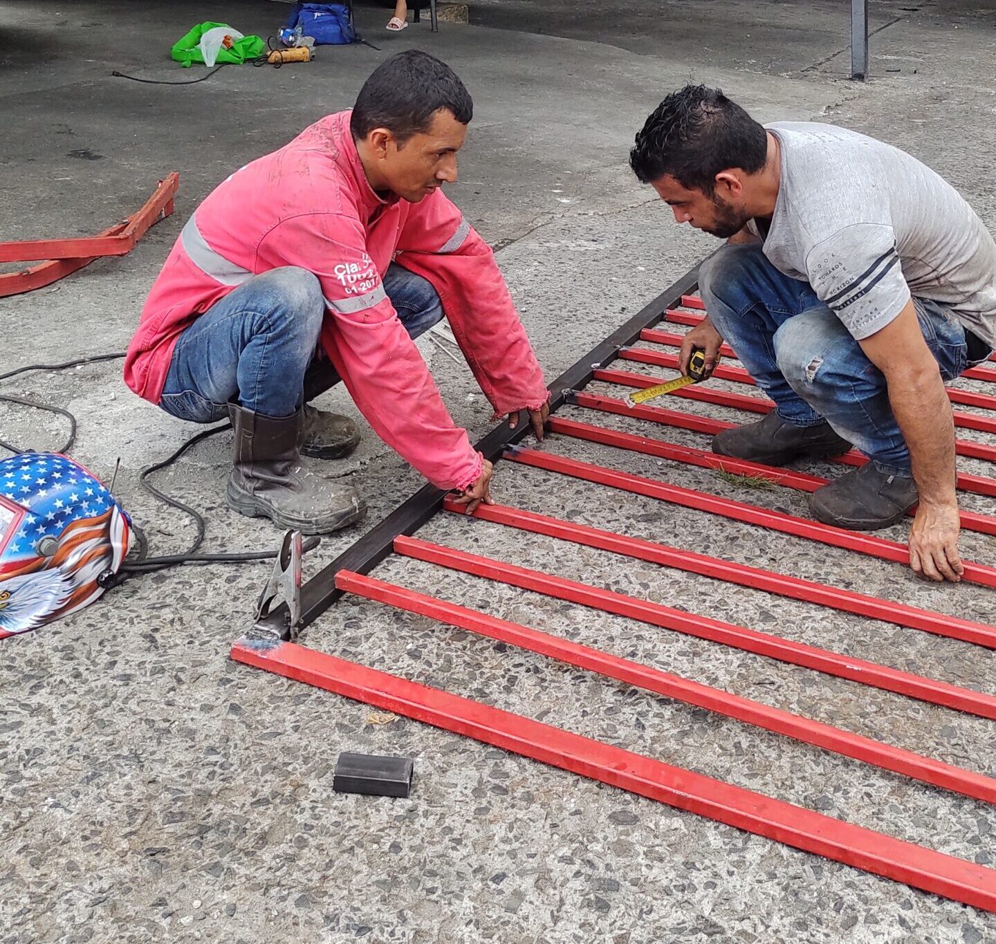 Dos hombres agachados sobre una estructura de portón de metal rectangular pintada de rojo en un suelo de concreto. Un hombre de rojo mide el centro mientras el otro sostiene una cinta métrica. Hay autos estacionados al fondo.