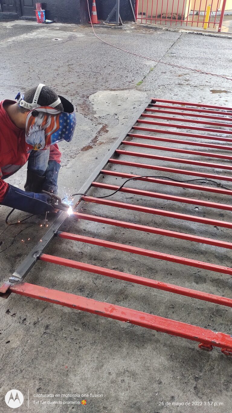 Un herrero con una máscara de soldar tricolor (con la bandera de EE. UU.) y ropa de trabajo roja está arrodillado en el suelo de concreto, soldando una unión en una esquina de una estructura de portón de metal rojo. Chispa brillante en el punto de soldadura.