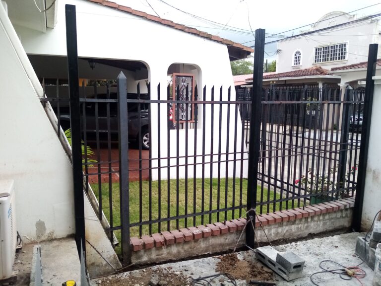 Vista desde el interior de un porche hacia el exterior a través de una reja de seguridad de metal negro. La reja tiene barrotes verticales con puntas afiladas y está montada sobre un murete bajo de ladrillos rojos. Los postes principales son vigas de acero negro robustas. En primer plano, hay materiales de construcción y herramientas en el suelo de concreto. Al fondo, la cochera de la casa y casas vecinas.