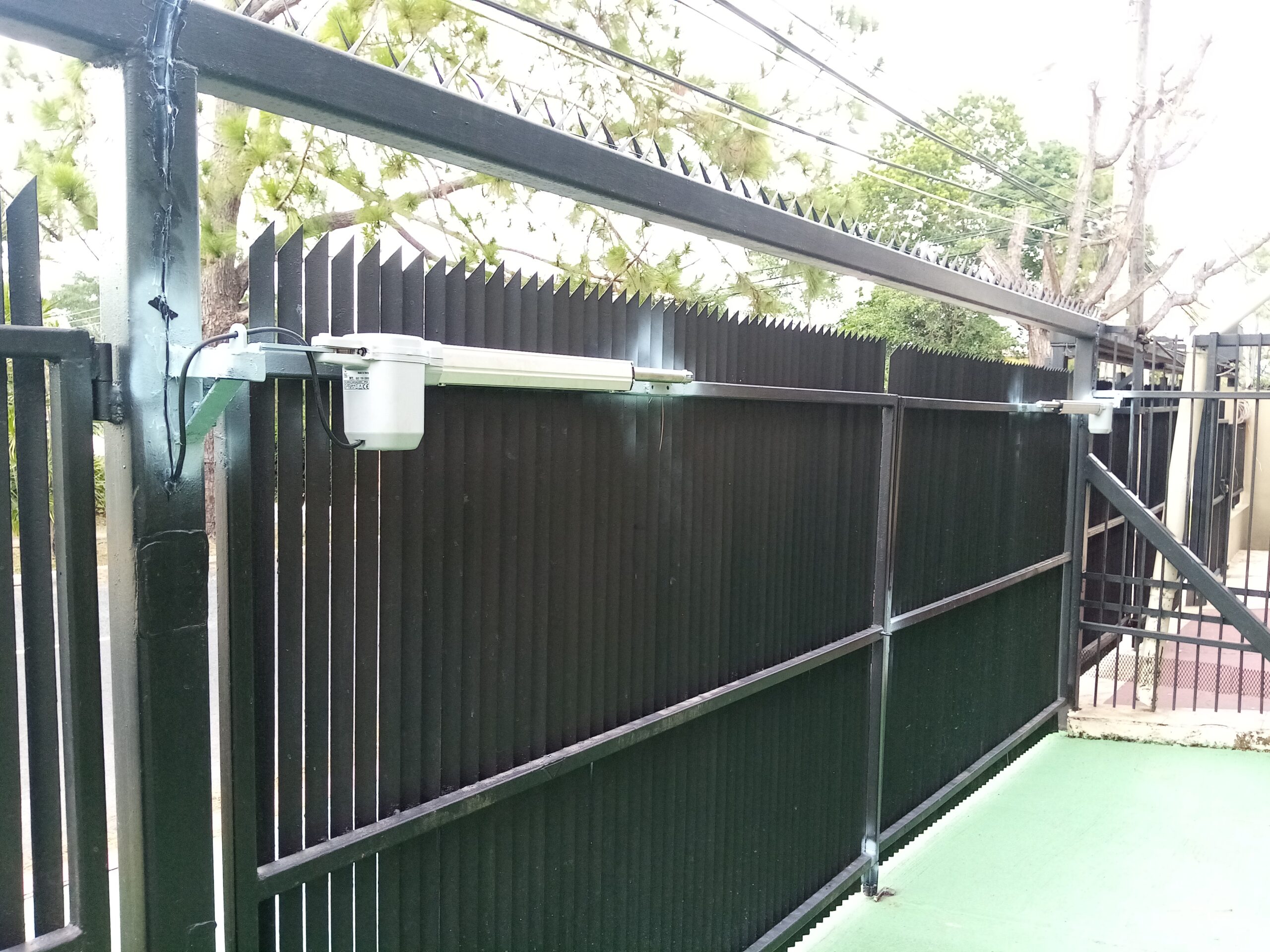Un motor eléctrico de color blanco y gris está montado en el poste superior izquierdo de una puerta metálica negra de barrotes verticales. La puerta está parcialmente visible, con una cerca de púas detrás y un fondo de árboles y cielo.