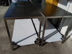 Dos mesas de trabajo largas hechas completamente de acero inoxidable con acabado brillante se encuentran dispuestas paralelamente sobre un suelo de concreto en el exterior. Cuentan con una repisa inferior plana cubierta con plástico protector blanco. En el fondo se aprecian cajas plásticas amarillas y una estructura blanca.