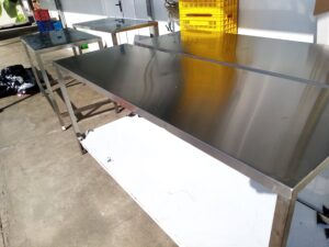 Varias mesas de trabajo de acero inoxidable de diferentes dimensiones están alineadas en un espacio exterior sobre pavimento de concreto. La superficie metálica refleja intensamente la luz del sol. Al fondo se observa una caja de plástico amarillo apilada y paredes blancas.