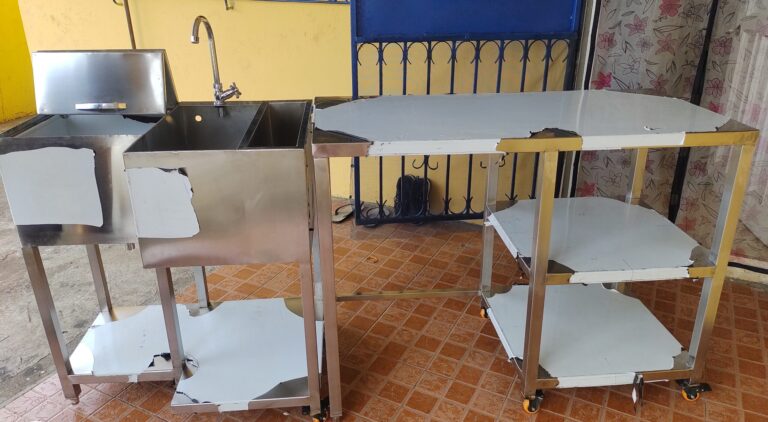 Conjunto de muebles auxiliares de acero inoxidable con fregadero, grifería tipo cuello de ganso y mesas con repisas inferiores sobre ruedas, ubicados en el exterior de una casa.