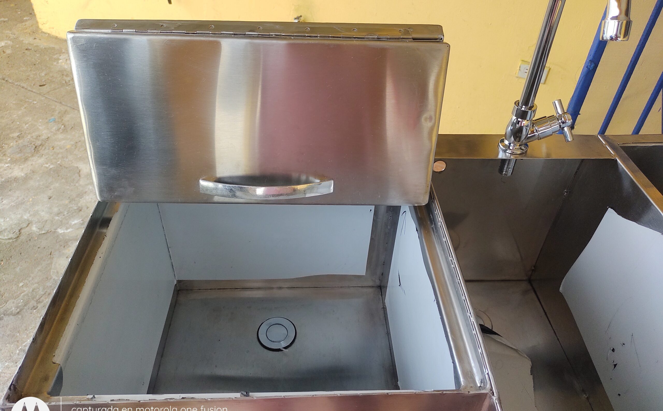 Vista de primer plano superior de una estación de acero inoxidable que muestra a la izquierda un compartimento profundo con su tapa metálica parcialmente abierta y asa, y a la derecha una cubeta de fregadero equipada con grifería tipo cuello de ganso.