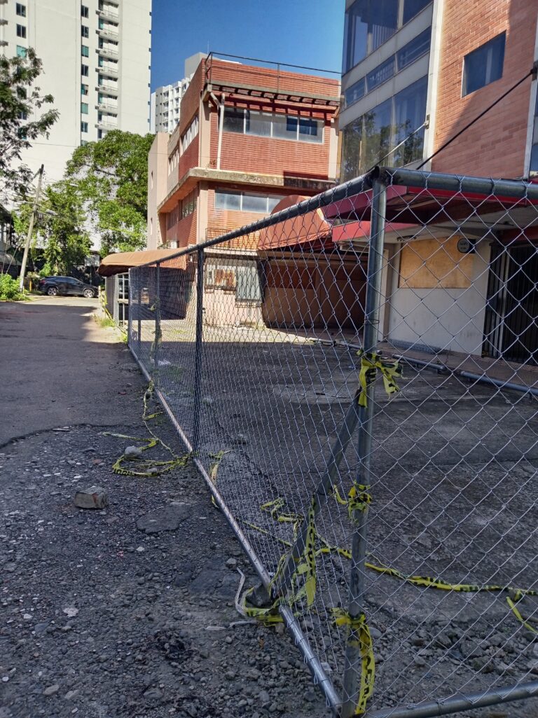 Una cerca de malla ciclónica delimita un área de estacionamiento de grava o tierra frente a un edificio de oficinas de dos plantas con fachada de ladrillo rojo y ventanas acristaladas. Cinta de precaución amarilla está atada a los postes de la cerca. Edificios altos al fondo.