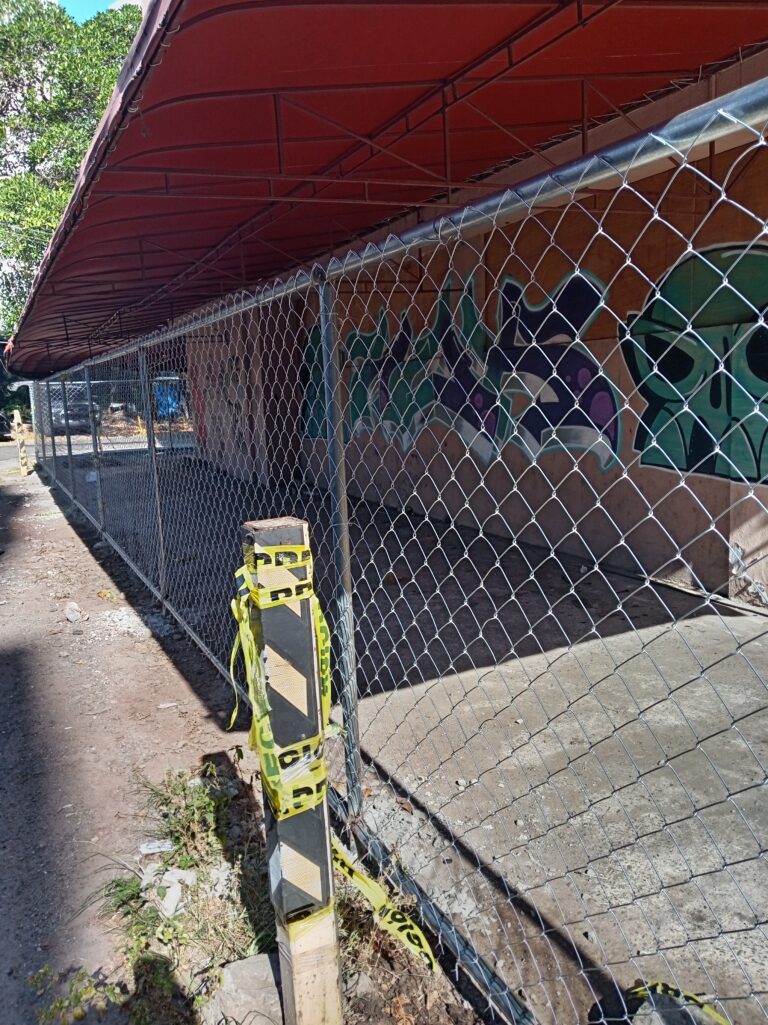 Una cerca de malla ciclónica se extiende en ángulo a lo largo de la acera. Detrás, la pared de un edificio de una sola planta está cubierta con coloridos graffitis. Un toldo rojo oscuro sobresale sobre la pared. Cinta de precaución amarilla está atada a un poste de la cerca en primer plano.