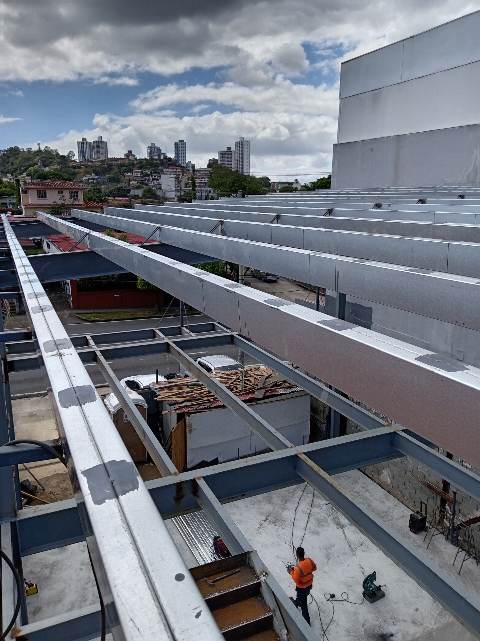 Vista superior de una obra en construcción con una serie de vigas y paneles metálicos entrelazados para el techo, con un trabajador abajo en el nivel inferior y un horizonte de ciudad al fondo bajo un cielo nublado.