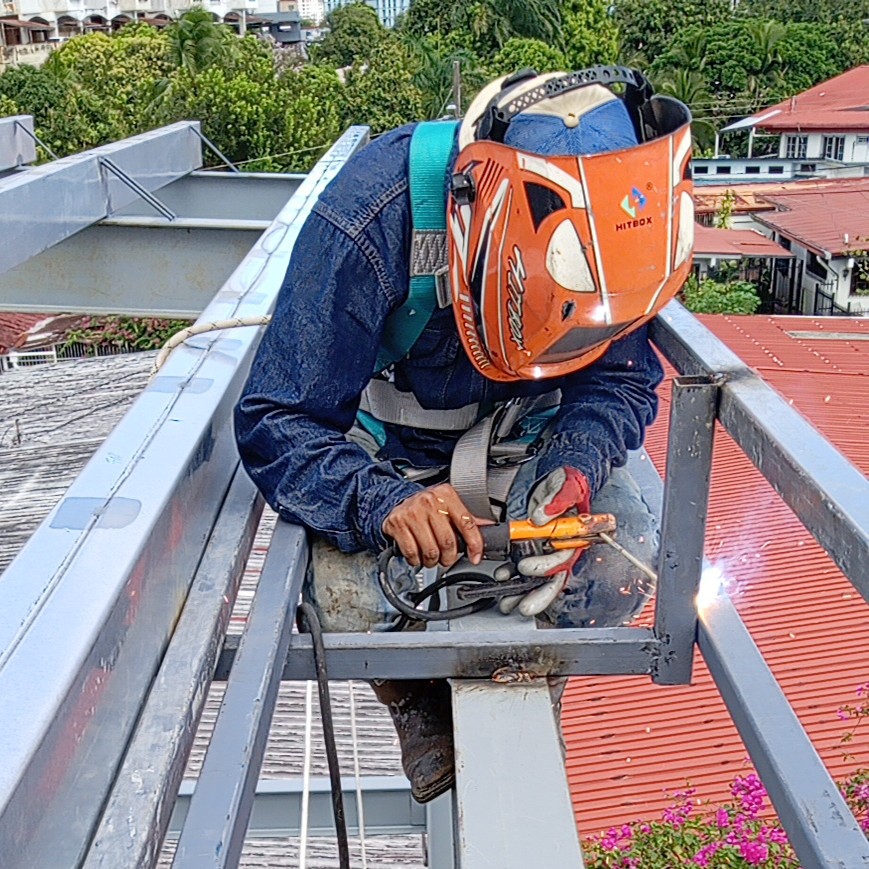 Un operario con equipo de protección, incluyendo arnés de seguridad y careta de soldar naranja, realiza trabajos de soldadura por arco sobre una estructura de acero galvanizado en el techo de un edificio.