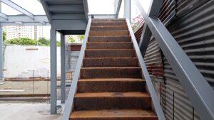 Primer plano de una escalera con peldaños de chapa estriada y con óxido superficial, flanqueada por vigas y soportes de acero pintados de gris en una obra en desarrollo.