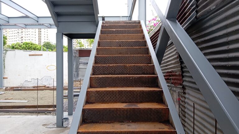 Primer plano de una escalera con peldaños de chapa estriada y con óxido superficial, flanqueada por vigas y soportes de acero pintados de gris en una obra en desarrollo.