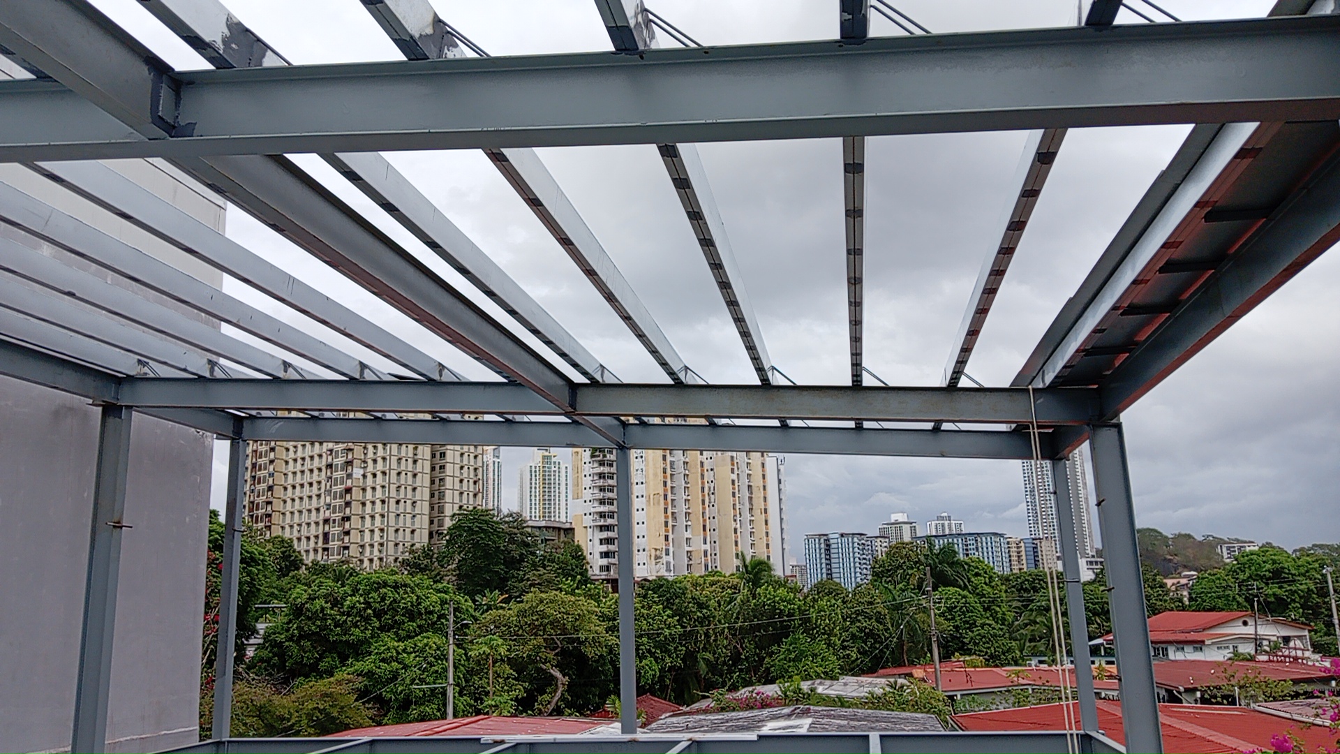 Vista desde una azotea en construcción, mirando hacia arriba y a través de una estructura de pérgola de acero gris y vigas perpendiculares, hacia un denso horizonte urbano con edificios altos y árboles bajo un cielo nublado.