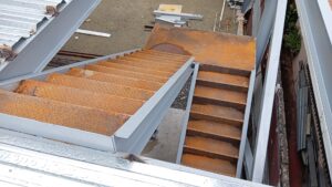 Vista de ángulo picado de una estructura de escaleras metálicas con peldaños de chapa estriada oxidada, vigas de soporte pintadas de gris y una cubierta de lámina ranurada en un área de obra.