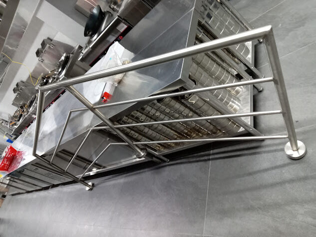 Una estructura tridimensional de tubos de acero inoxidable con diseño geométrico y angular se encuentra en primer plano sobre un piso gris, con mesas de trabajo de cocina industrial al fondo.