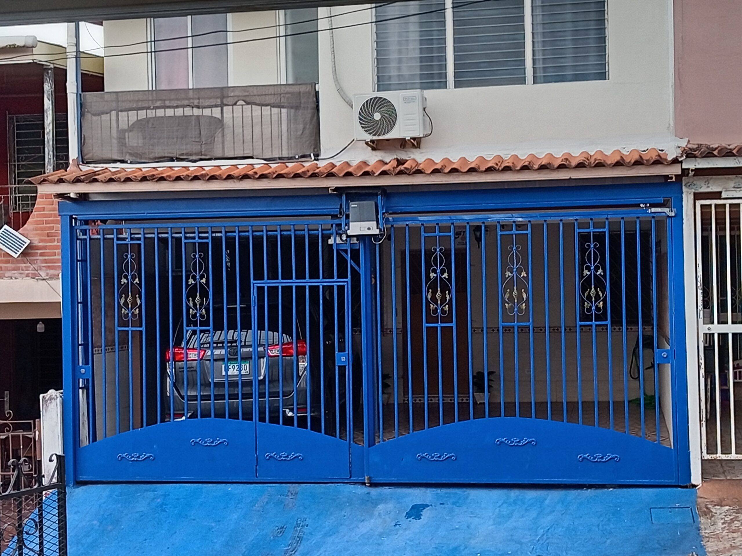 Un portón de garaje de metal pintado de azul brillante con un diseño ornamental curvado en la parte inferior y paneles de rejas con detalles dorados está cerrado. Se ve parcialmente un vehículo gris oscuro detrás de la puerta de la reja. Un edificio de dos plantas con techo de tejas y una unidad de aire acondicionado está detrás.