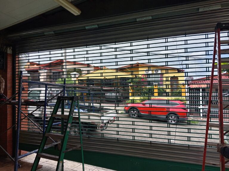 Vista desde el interior de un taller o garaje hacia el exterior a través de una persiana de seguridad enrollable de color verde parcialmente abierta. Se ven escaleras de mano (una verde, una roja) y andamios a los lados. Afuera, hay una camioneta blanca estacionada y un SUV rojo, detrás de casas suburbanas.