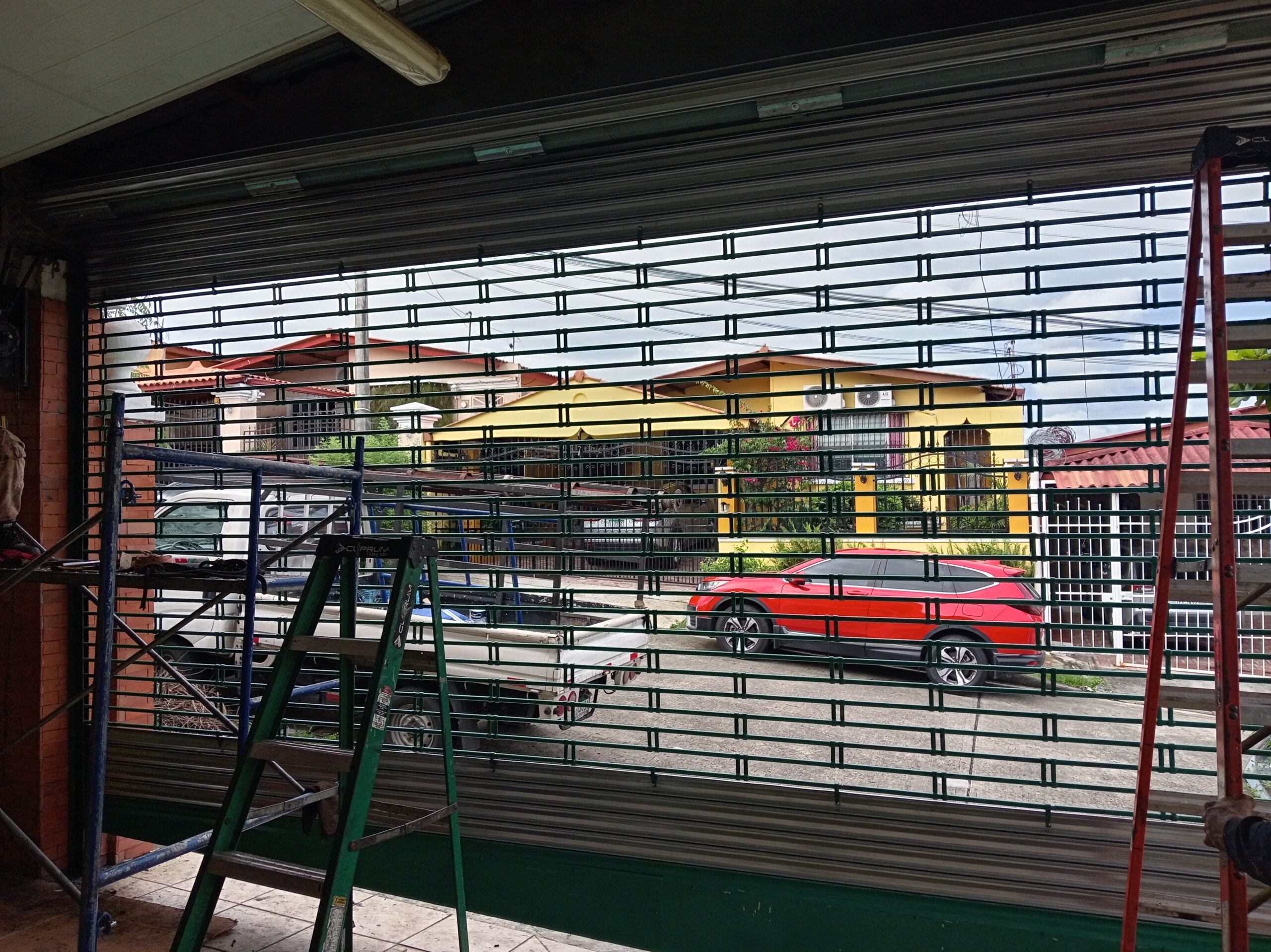 Vista desde el interior de un taller o garaje hacia el exterior a través de una persiana de seguridad enrollable de color verde parcialmente abierta. Se ven escaleras de mano (una verde, una roja) y andamios a los lados. Afuera, hay una camioneta blanca estacionada y un SUV rojo, detrás de casas suburbanas.