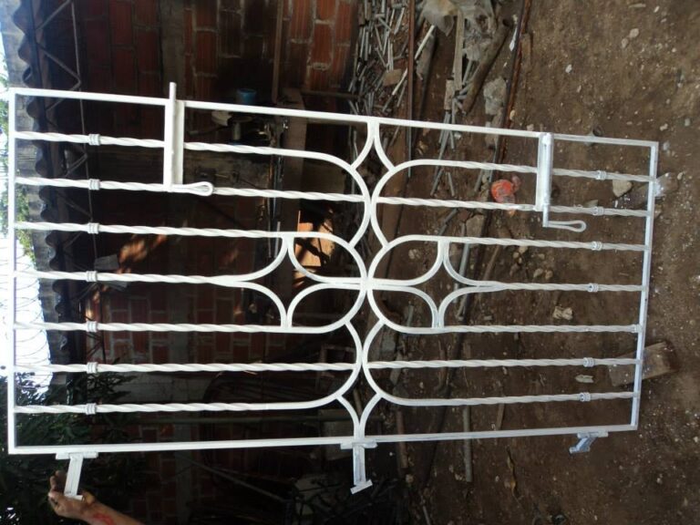 Una reja de seguridad para ventana de metal blanco, con un diseño ornamental complejo que incluye volutas y formas geométricas romboidales en el centro, reposa en el suelo de tierra de un taller de herrería. El fondo es un muro de bloques de ladrillo visto y escombros.