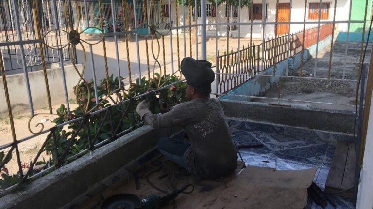 Un herrero, visto por detrás y vestido con ropa de trabajo y una máscara de soldar negra, está agachado en el balcón de una casa, realizando trabajos de soldadura en una reja metálica. Alrededor hay herramientas eléctricas y la reja está junto a un seto. En el fondo, casas de vecindario.