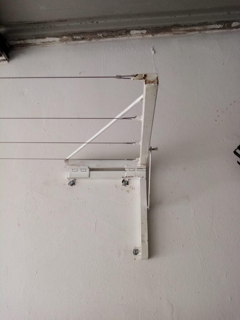 Soporte de pared de metal blanco plegable para tendedero, con cables de acero, montado en una pared de estuco blanco en Panamá. Servicio de instalación y mantenimiento.