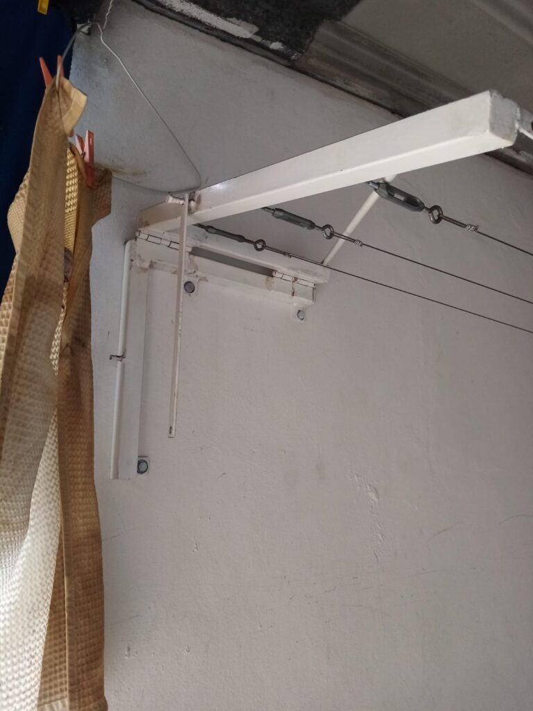 Vista en ángulo de un soporte de pared metálico blanco para tendedero con tensores de cable de acero, instalado en una pared de estuco en Panamá. Reparación y fabricación a medida.