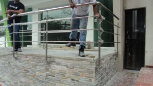 Dos obreros trabajan en la instalación de una barandilla de acero inoxidable compuesta por un pasamanos y tubos horizontales paralelos en la entrada elevada de un edificio con revestimiento de piedra.