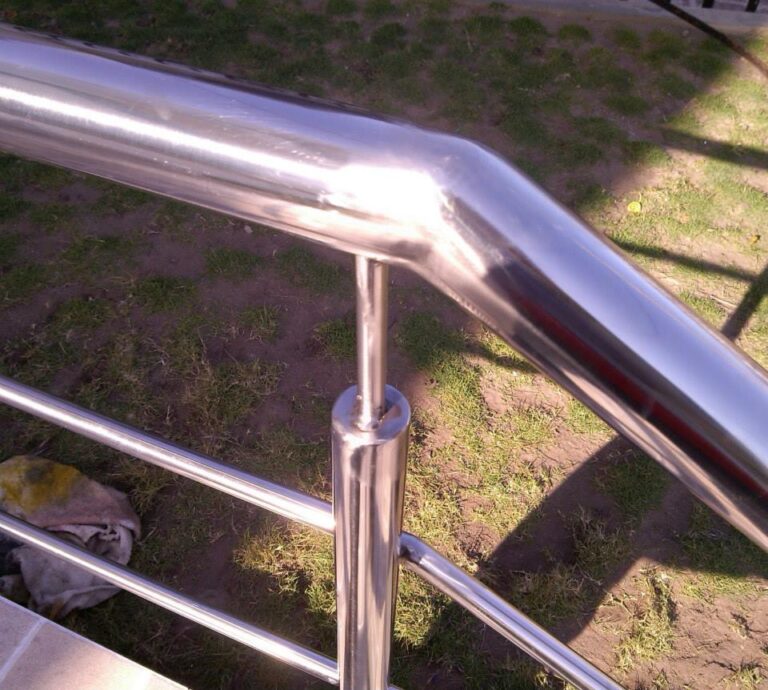 Primer plano de la unión curvada y soldada de un pasamanos tubular de acero inoxidable con un poste de soporte vertical en un entorno exterior con césped de fondo.