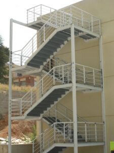 Vista exterior de una edificación con una escalera metálica de varios niveles, estructura de soporte blanca, peldaños oscuros y barandas de seguridad con barras horizontales.